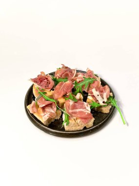 Seramik tabakta servis edilen prosciutto, roka ve yumuşak peynirli taze bruschetta aperatifi. Beyaz arka planda minimalist kompozisyon, restoran menüsü ya da mutfak içeriği için mükemmel.