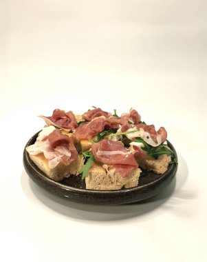 Seramik tabakta servis edilen prosciutto, roka ve yumuşak peynirli taze bruschetta aperatifi. Beyaz arka planda minimalist kompozisyon, restoran menüsü ya da mutfak içeriği için mükemmel.