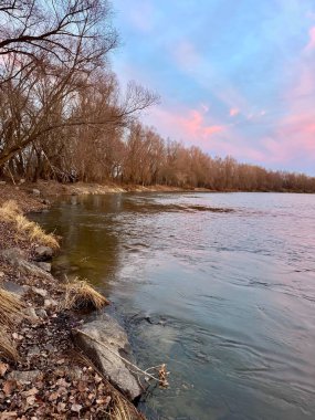 Nehrin üzerinde gün batımı, sudaki gökyüzünün renklerinin yansımasıyla. Ön planda kuru çimenler ve kıyıda taşlar var. Ufukta resim şeklinde siluet oluşturan ağaçlar var.