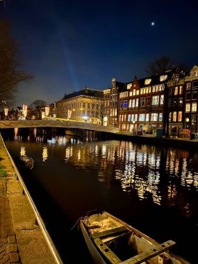 Geceleri Amsterdam: yansıyan ışıkları, eski evleri, aydınlatılmış köprüsü olan kanal. Ön planda kıyıya yakın eski ahşap bir tekne var, atmosfer ekliyor..