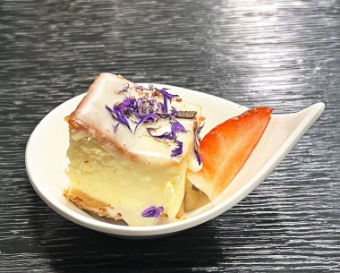 Kurutulmuş menekşeler ve gümüş taneciklerle süslenmiş, beyaz bir tabakta bir dilim elmayla servis edilen nefis bir tatlı. Siyah arkaplan yemeğin dokusunu ve detaylarını vurgular.