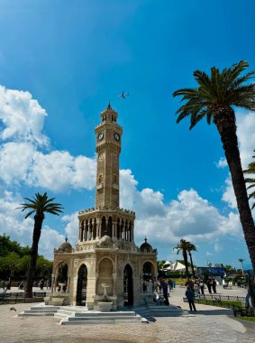 İzmir, Türkiye 19 Nisan 2024. Osmanlı döneminin simgelerinden Konak Meydanı 'ndaki tarihi İzmir Saat Kulesi. Güneşli bir günde tepemizde bir uçak uçarken palmiye ağaçları kuleyi çerçeveler..