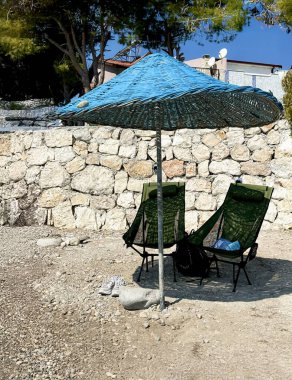Blue wicker sun umbrella on a stone wall background, two chairs for relaxation nearby. Summer mood, atmosphere of relaxation and outdoor recreation.
