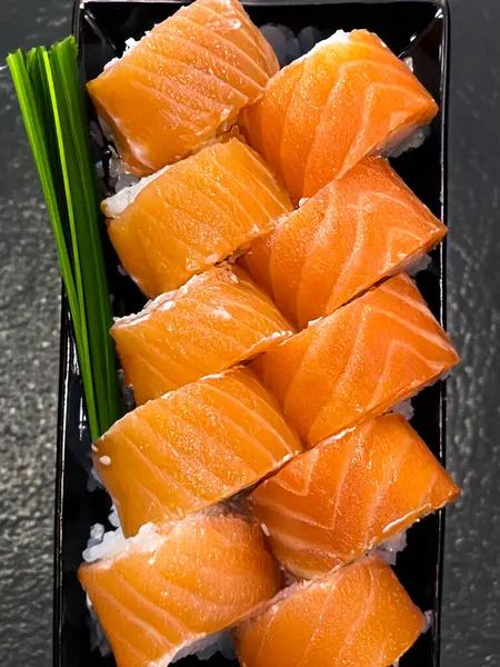 Siyah tabakta somonla birlikte suşi yuvarlanıyor. Taze somon filetosu pirinç rulolarını eşit şekilde kaplar ve mükemmel bir tat dengesi yaratır..