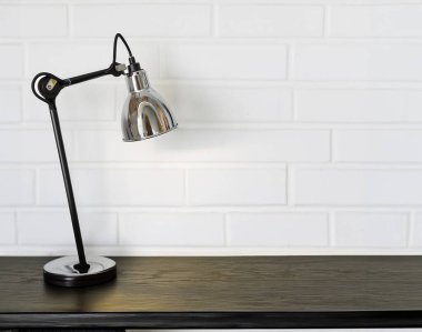 Beyaz tuğla duvara karşı metal abajurlu modern masa lambası. Çalışma alanının minimalist iç ve rahat atmosferi.