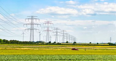 Elektrik hatları ve rüzgar türbinleri mavi gökyüzünün altındaki yeşil alanlar boyunca uzanıyor. Altyapı, yenilenebilir enerji ve kırsal kalkınma kavramı.