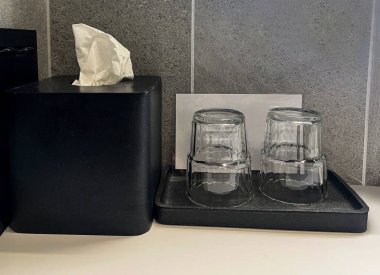 Two empty glass cups placed upside down on a tray next to a black tissue box in a modern bathroom interior.