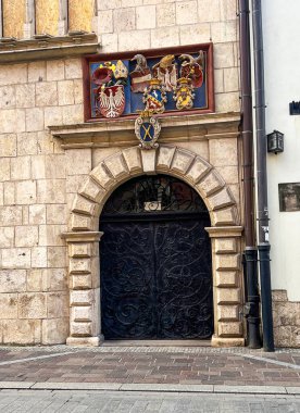 Editorial image of a historic stone building facade with an arched black wrought iron gate and colorful heraldic coats of arms above the entrance. Captured on October 3, 2025, in Krakow, Poland.