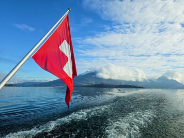 İsviçre dağ manzaralı, hareket eden bir teknenin arkasından çekilmiş. Rüzgarda dalgalanan İsviçre bayrağı
