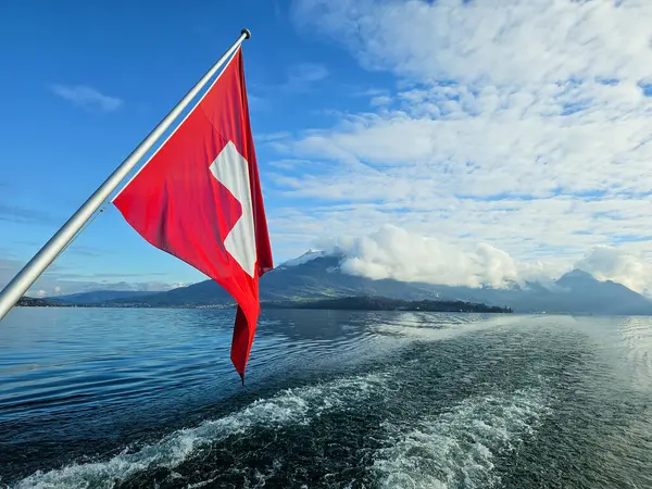 İsviçre dağ manzaralı, hareket eden bir teknenin arkasından çekilmiş. Rüzgarda dalgalanan İsviçre bayrağı