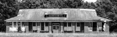 The black-and-white photo captures an old, abandoned schoolhouse, its facade a mix of weathered brick and decaying wood. The building exudes an air of neglect, with broken windows and a sagging roofline that speak of years of disuse. 