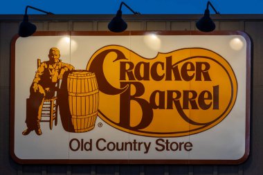 Görüntü, bir binanın dış duvarına asılmış orijinal Cracker Barrel Old Country Store logosunu gösteriyor. Dikdörtgen tabelanın koyu kahverengi kenarlı krem renkli bir arka planı var. Logonun sol tarafında eski kanepe giymiş bir adam var.
