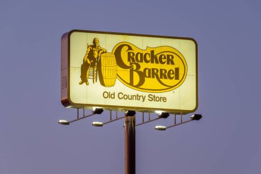 Görüntü, bir binanın dış duvarına asılmış orijinal Cracker Barrel Old Country Store logosunu gösteriyor. Dikdörtgen tabelanın koyu kahverengi kenarlı krem renkli bir arka planı var. Logonun sol tarafında eski kanepe giymiş bir adam var.