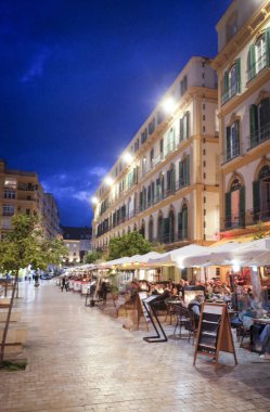 Malaga, İspanya. 7 Mart 2025. Gün batımında Mlaga 'daki Plaza de la Merced' in atmosferik manzarası ılık aydınlatma, hareketli kafeler ve zengin İspanyol kültürü. (Tanınabilir kişiler ve marka isimleri dijital olarak kaldırıldı)