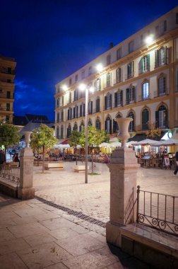 Malaga, İspanya. 7 Mart 2025. Gün batımında Mlaga 'daki Plaza de la Merced' in atmosferik manzarası ılık aydınlatma, hareketli kafeler ve zengin İspanyol kültürü. (Tanınabilir kişiler ve marka isimleri dijital olarak kaldırıldı)