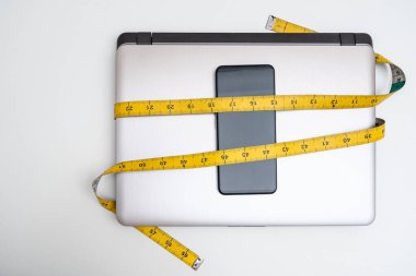 Bilgisayar ve akıllı telefon beyaz arka planda mezura ile bağlanmış. Dijital gecikme, sosyal medya aşırı kullanımı ve iş hayatı dengesizliği kavramları