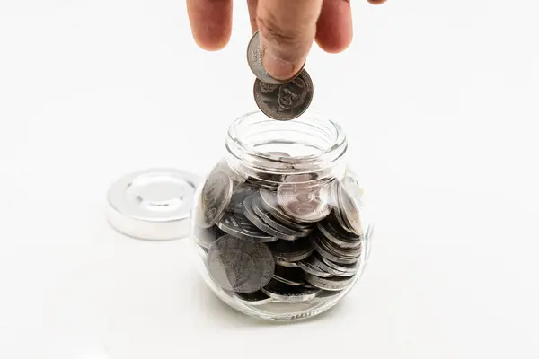 Bozuk paraları beyaz arka plandaki küçük bir kavanoza koyan bir el. Tasarruf, bütçe veya mali planlama kavramları