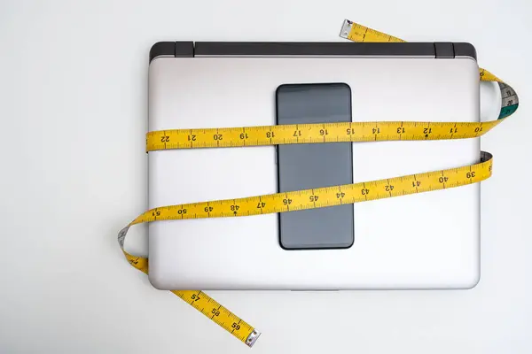 Bilgisayar ve akıllı telefon beyaz arka planda mezura ile bağlanmış. Dijital gecikme, sosyal medya aşırı kullanımı ve iş hayatı dengesizliği kavramları