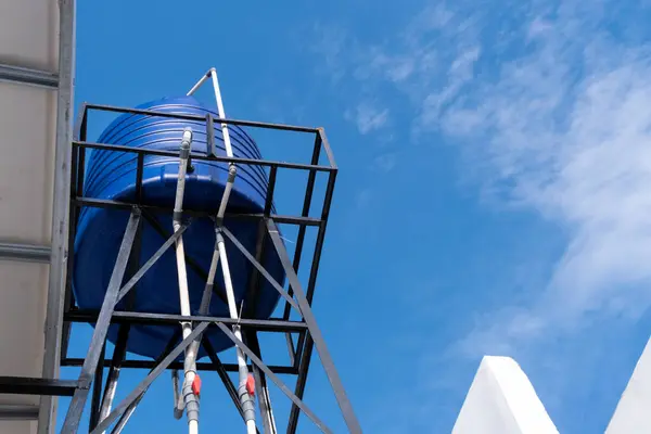 Uzun bir çatı su tankı bir evin üzerinde duruyor, parlak mavi bir gökyüzüne karşı.