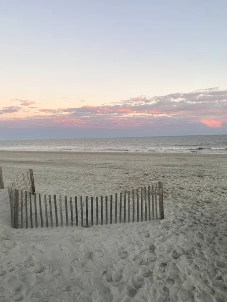 Gün batımında kumlu bir plajda, üzerinde pembe bulutlar olan çitlerle çevrili bir bölge..