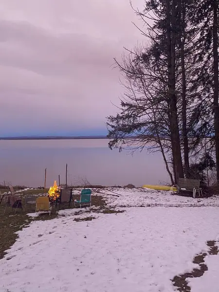 Kamp ateşi, sandalyeler, ağaçlar ve pembe gökyüzü olan karlı bir göl manzarası..