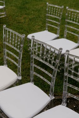 Her biri yumuşak beyaz yastıklarla donatılmış şeffaf sandalyeler, dışarıdaki yemyeşil çimlerin üzerine zarif bir şekilde yerleştirilir.