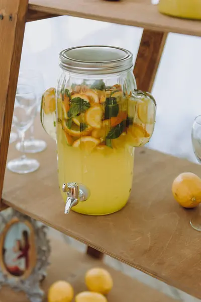 İçi ferahlatıcı limonata dolu cam bir kavanoz şu anda olgunlaşmış limonların yanındaki ahşap bir rafta duruyor.