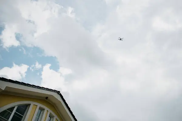 Bir dron zarif bir şekilde açık mavi gökyüzünde uçuyor. Altında huzur içinde duran büyüleyici sarı bir evin üzerinde.