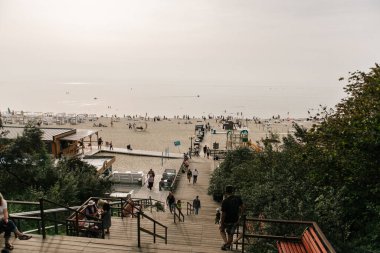 Bulanık bir görüntü, güneşli bir sahile doğru giden merdivenlerden inen bir grup bireyi yakalar.