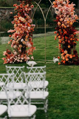 Güzel bir ortamda, zarif beyaz sandalyeler titizlikle düzenlenmiştir çarpıcı bir çiçek kemerinin önünde, canlı çiçeklerle süslenmiş.