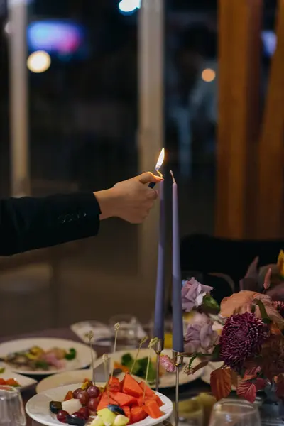 Bir insan lezzetli yiyecekler ve diğer şeylerle süslenmiş bir masaya konmuş bir mumu dikkatle yakıyor.