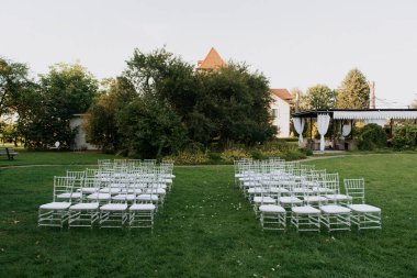 Resimli bir çayır, düzenli bir sıra el değmemiş beyaz sandalyeler dikkatlice dizilir, açık hava etkinliği için davetkar bir atmosfer yaratır.