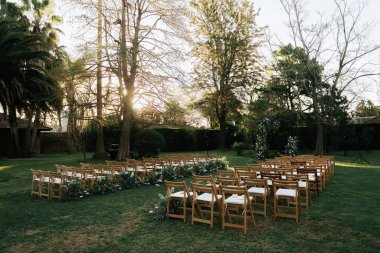 Bir sıra güzel işlenmiş ahşap sandalyeler canlı doğa ve manzarayla çevrili yemyeşil bir alanda düzgünce dizilmiştir.