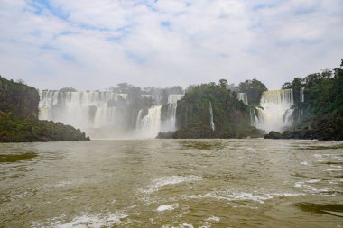 Bu görkemli sahne, arka planda büyüleyici bir şelale ile tamamlanmış, sakin bir nehrin akışını içerirken, yemyeşil ağaçlar ön planda gururla durur.