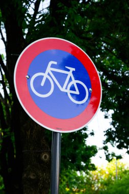 Çarpıcı bir kırmızı ve mavi tabela belirgin bir şekilde gösterilir üzerinde bisiklet güvenliğinin sembolü olan bir bisiklet resmedilir.