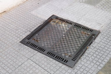 Sağlam bir rögar kapağı fayansla döşenmiş bir kaldırıma yerleştirilmiş, onu çevreleyen kentsel zemin ortamına kusursuz bir şekilde uyum sağlıyor.