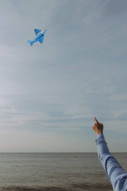 Bir insan coşkuyla gökyüzünde süzülen parlak mavi kağıt bir uçağı işaret ediyor, o anın güzelliğini yakalıyor.