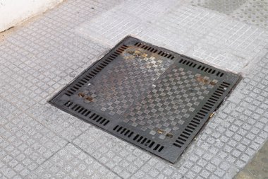 Sağlam bir rögar kapağı fayansla döşenmiş bir kaldırıma yerleştirilmiş, onu çevreleyen kentsel zemin ortamına kusursuz bir şekilde uyum sağlıyor.