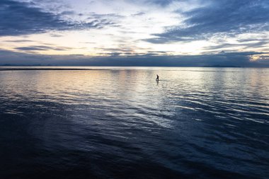 Gün batımında deniz kenarında bir plajda çekim ve sığ, pürüzsüz yansıtan suda duran bir kişi.