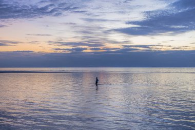 Gün batımında deniz kenarında bir plajda çekim ve sığ, pürüzsüz yansıtan suda duran bir kişi.