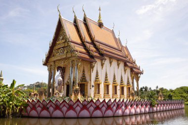 Ornate Thai temple surrounded by water, showcasing intricate golden decorations, vibrant colors, and traditional architecture against a clear sky. Highlights the beauty of Thai spiritual heritage. clipart