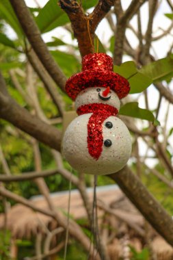 Festive Christmas snowman ornament with a glittery golden hat hanging on a tree branch outdoors. Perfect for holiday decorations, nature-inspired celebrations, and winter themes. clipart