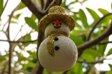 Festive Christmas snowman ornament with a glittery golden hat hanging on a tree branch outdoors. Perfect for holiday decorations, nature-inspired celebrations, and winter themes. clipart