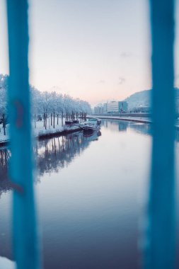 River with a Boat Docked to the Snow Coverd Shore in the City Viewed Through the Railings of a Bridge on a Cold Winter Day clipart