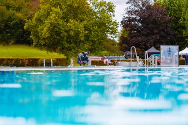 Parlak mavi bir yüzme havuzuna enerjik bir şekilde atlayan bir köpeğin canlı görüntüsü yemyeşil ve huzurlu bir park düzeniyle çevrili..
