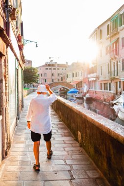 Yaşlı bir adam gün batımında Venedik, İtalya 'da büyüleyici bir kanalda geziniyor. Etrafı suyla çevrili ve sıcak güneş ışığıyla yıkanmış tarihi mimariyle rahat rahat yürüyor..