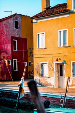 Canlı sarı ve kırmızı Venedik evleri sakin bir kanal boyunca uzanır. Sıcak öğleden sonra güneşinin altında Burano adasında kırsal dokular ve huzurlu yansımalar sergiler..