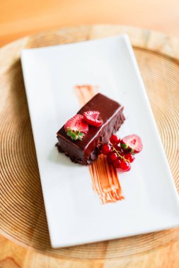 Lezzetli çikolatalı tatlı, taze çilekler ve kırmızı üzümlerle süslenmiş, beyaz bir tabakta sunuluyor, gurme yemekleri için cazip ve sofistike bir gastronomik zevk sunuyor.