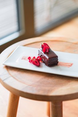 Taze ahududu ve kuşüzümü ile süslenmiş zengin bir çikolatalı pasta dilimi, kırsal ahşap bir masanın üzerinde zarif bir şekilde sunulur, hoşgörü ve gurme yayılır..