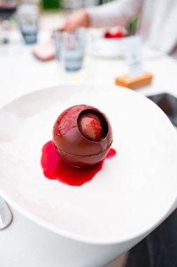 Close-up of an exquisite chocolate dessert sphere filled with decadent mousse, draped in vibrant raspberry sauce. The elegant presentation creates a sense of indulgence and culinary artistry. clipart